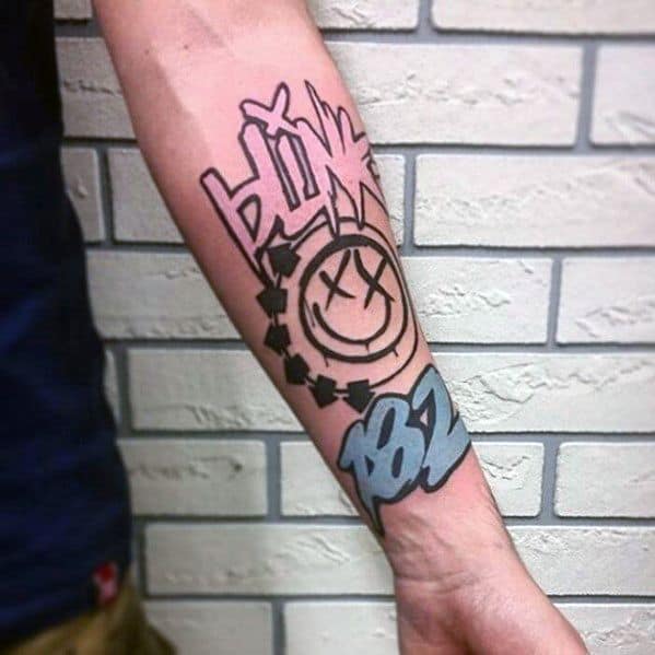 Tattoo of the Blink-182 smiley face logo with a stylized "BLINK" text and "182" in bold, colorful lettering, placed on the forearm.