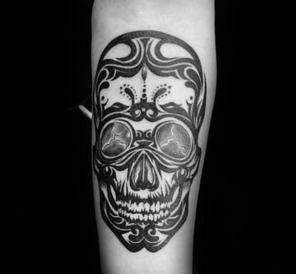 Detailed tribal skull tattoo on forearm with bold patterns, ornamental swirls, and circular mirrored eyes for a dramatic, artistic effect.