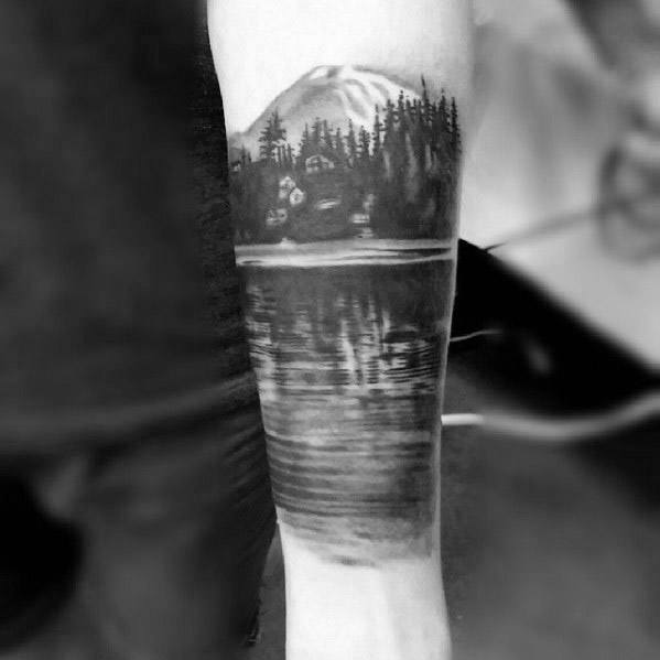 Black and grey forearm tattoo showing a mountain cabin, tall pines, and calm lake with detailed reflections for a peaceful nature scene.