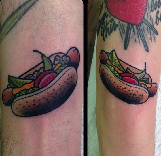 Hot dog tattoo featuring a sausage with mustard, ketchup, relish, onions, and tomatoes in a poppy seed bun, on the forearm.