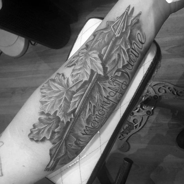Black and grey forearm tattoo of an arrow surrounded by maple leaves with the script “In memory of Duane” wrapped around the shaft.