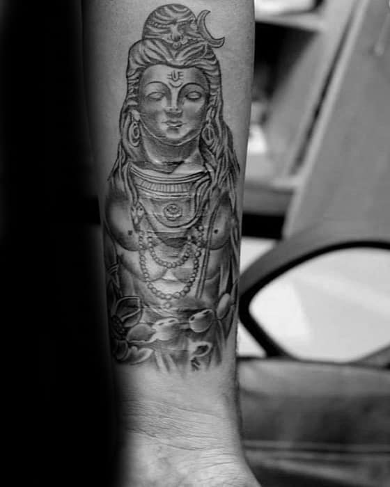 Forearm tattoo of Lord Shiva with closed eyes, adorned with rudraksha beads, crescent moon, and lotus flowers at the base in fine black shading.
