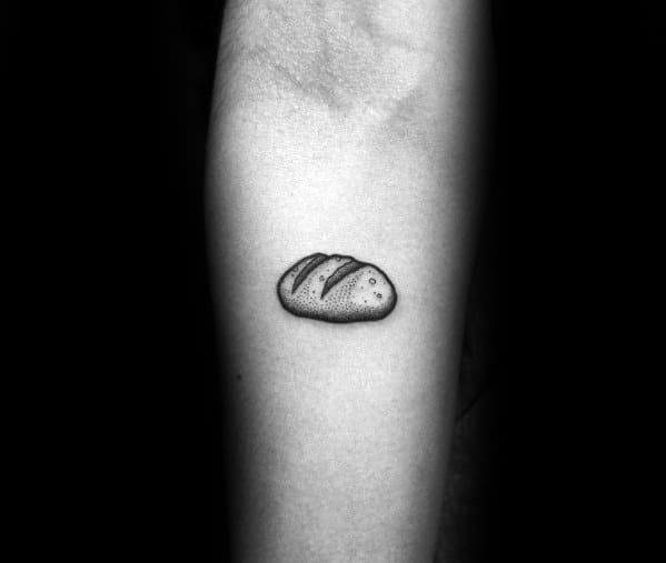 Small black ink tattoo of a rustic bread loaf with fine shading and simple details, creating a minimalist yet textured look on the forearm.