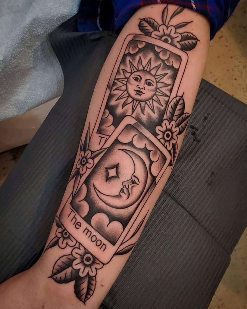Bold black ink tattoo featuring sun and moon tarot cards, adorned with floral details and shading, creating a mystical and celestial design.