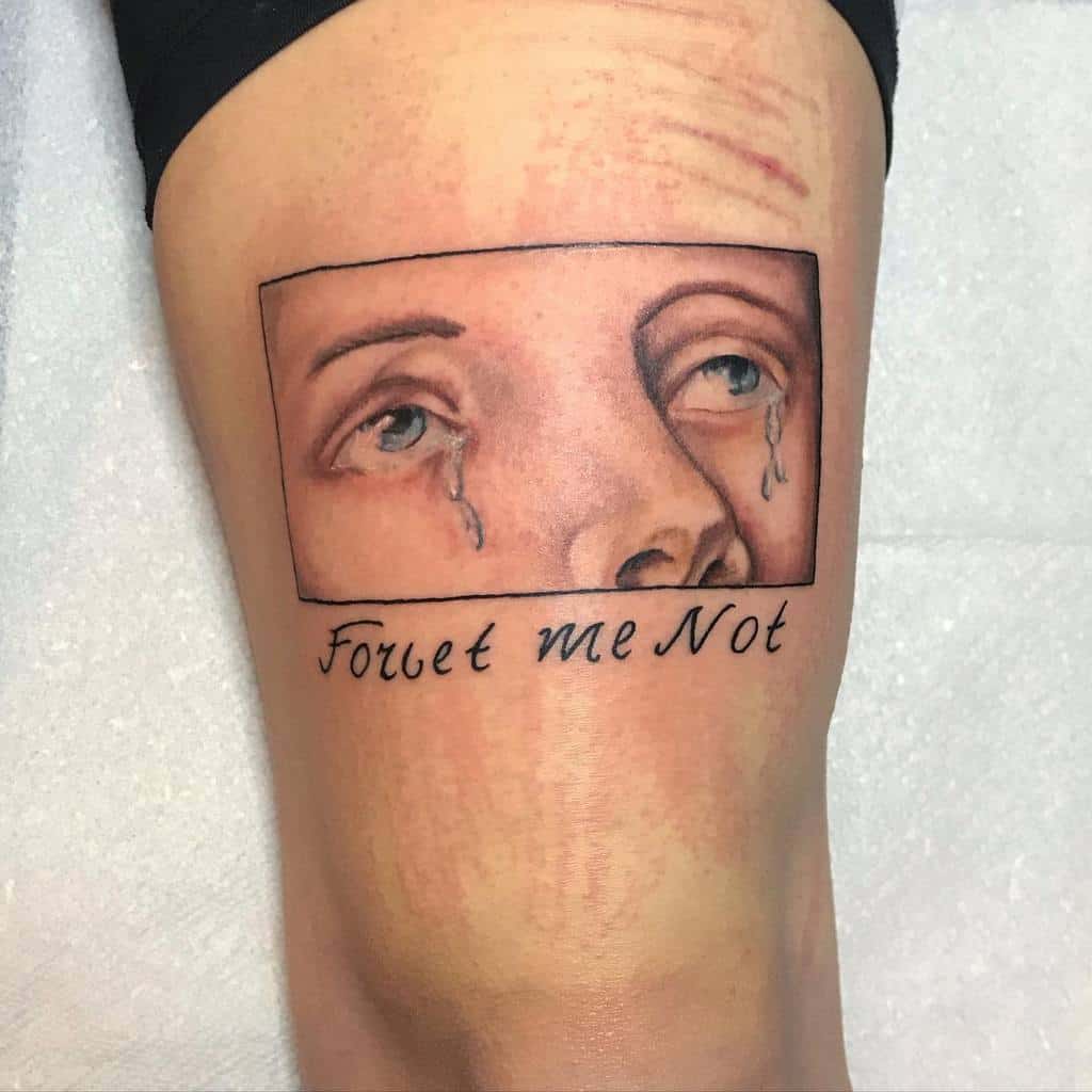 Tattoo of a somber pair of eyes with tears inside a rectangle, below the words "Forget me Not" are inked