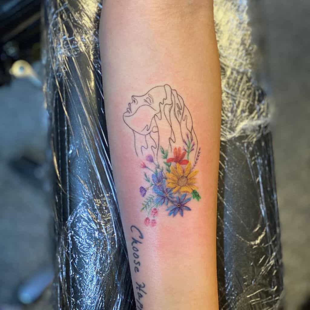 Elegant fine-line tattoo of a woman's face in profile with flowing hair, surrounded by vibrant watercolor-style flowers in shades of yellow, blue, and red