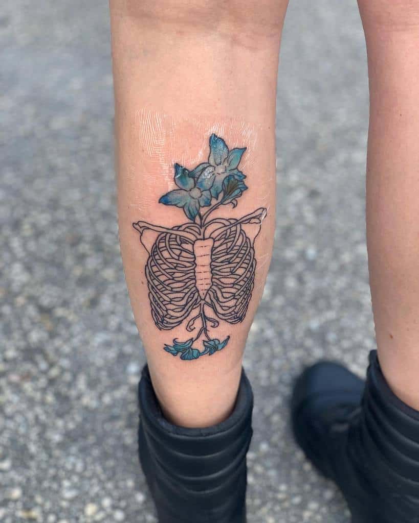 Fine-line tattoo of a ribcage with blue flowers growing from within, symbolizing life and transformation