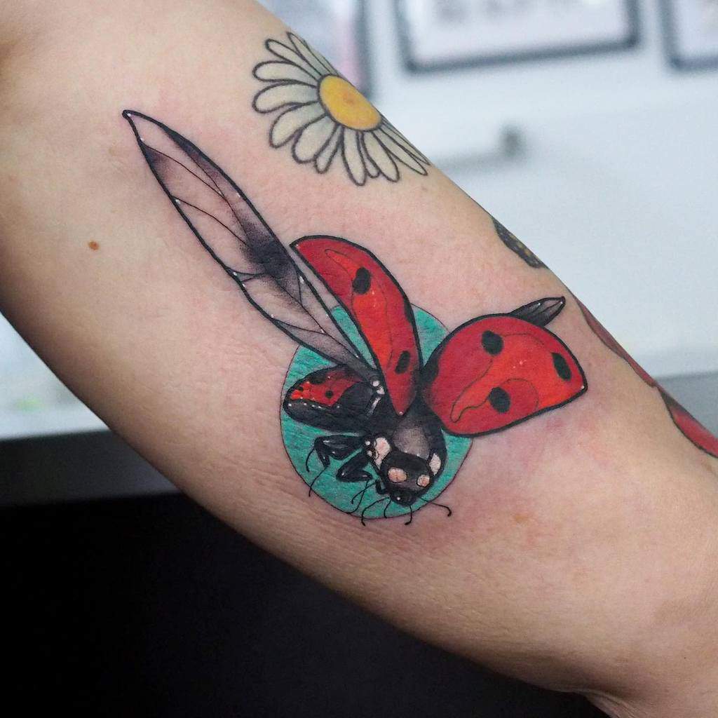 Neo-traditional ladybug tattoo in flight with bold red wings, fine black details, and a teal background, paired with a delicate daisy above.