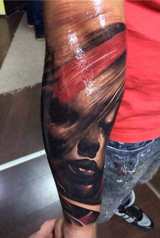 Hyper-realistic tattoo of a woman's face with a red streak across her eyes on an arm