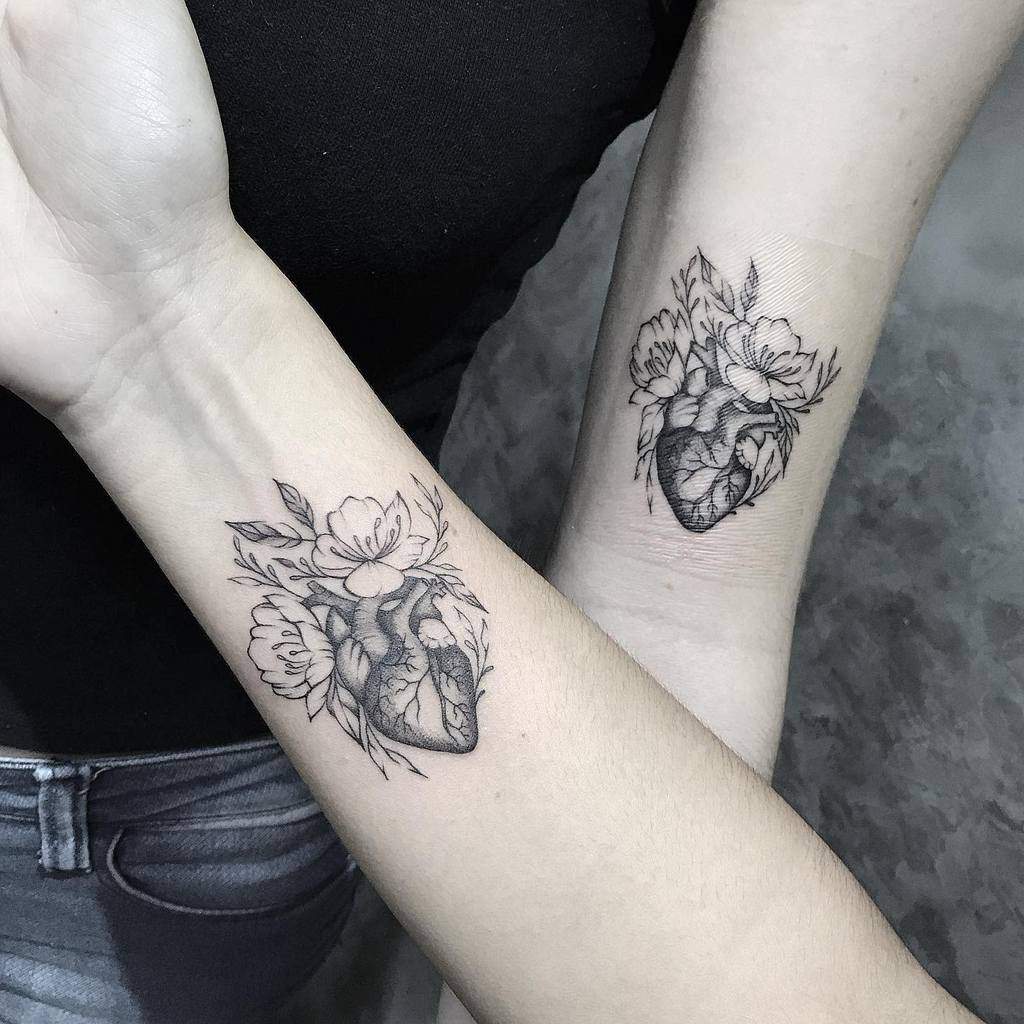 Matching sister tattoos featuring an anatomical heart intertwined with delicate flowers and leaves, symbolizing love and connection
