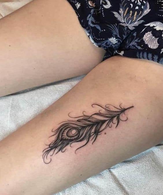 Elegant black ink peacock feather tattoo on the thigh, featuring delicate linework, fine shading, and a detailed eye pattern.