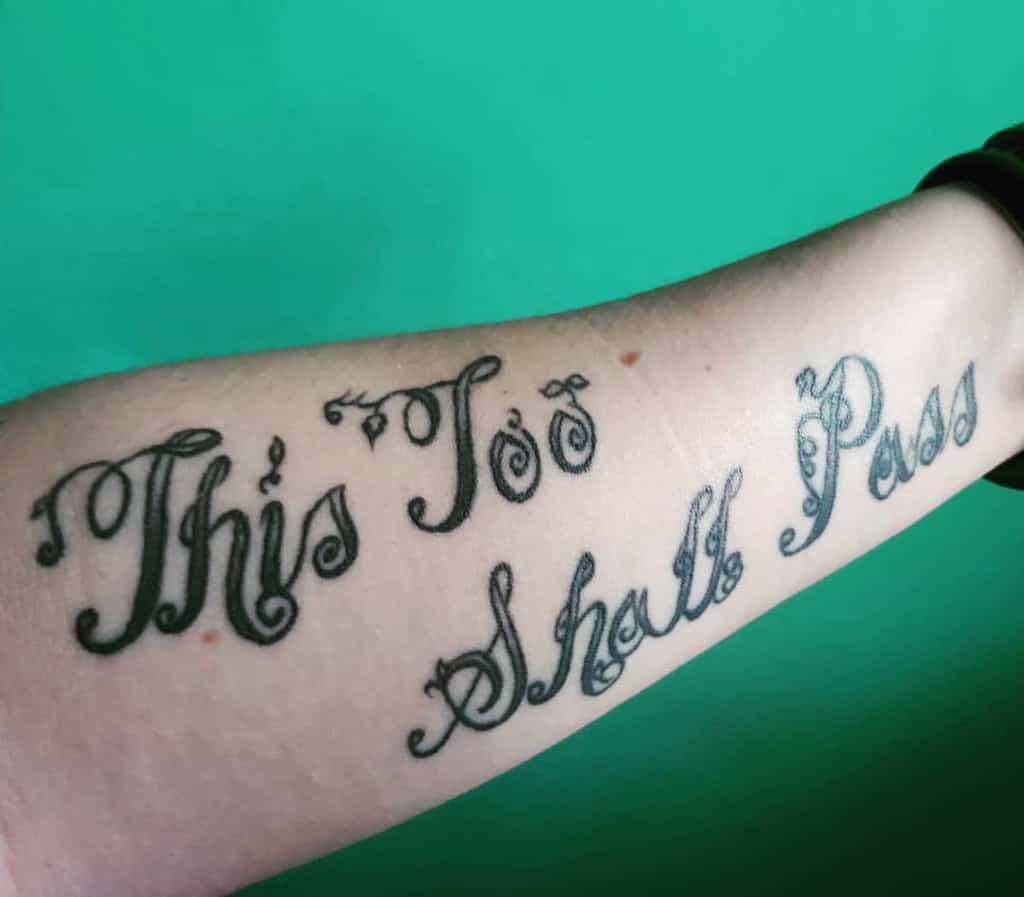 A bold "This Too Shall Pass" tattoo with an ornate, decorative font. The elaborate lettering gives it a unique and eye-catching aesthetic, emphasizing the phrase’s deep meaning of resilience and impermanence.