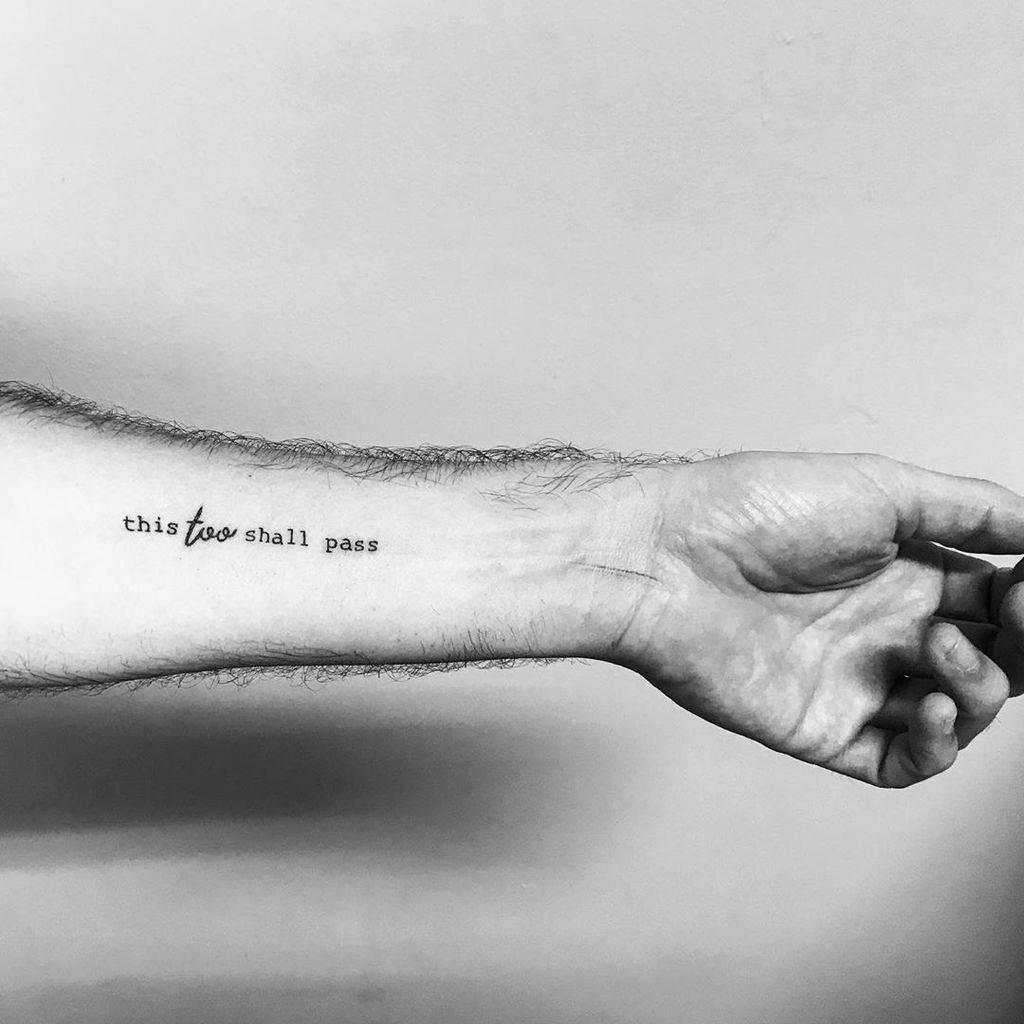 This image showcases a tattoo on the forearm, featuring the phrase "this too shall pass" in lowercase lettering with an emphasis on the word "too" in a slightly different font.