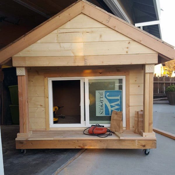 A wooden playhouse with white-framed windows and a pitched roof, set on wheels
