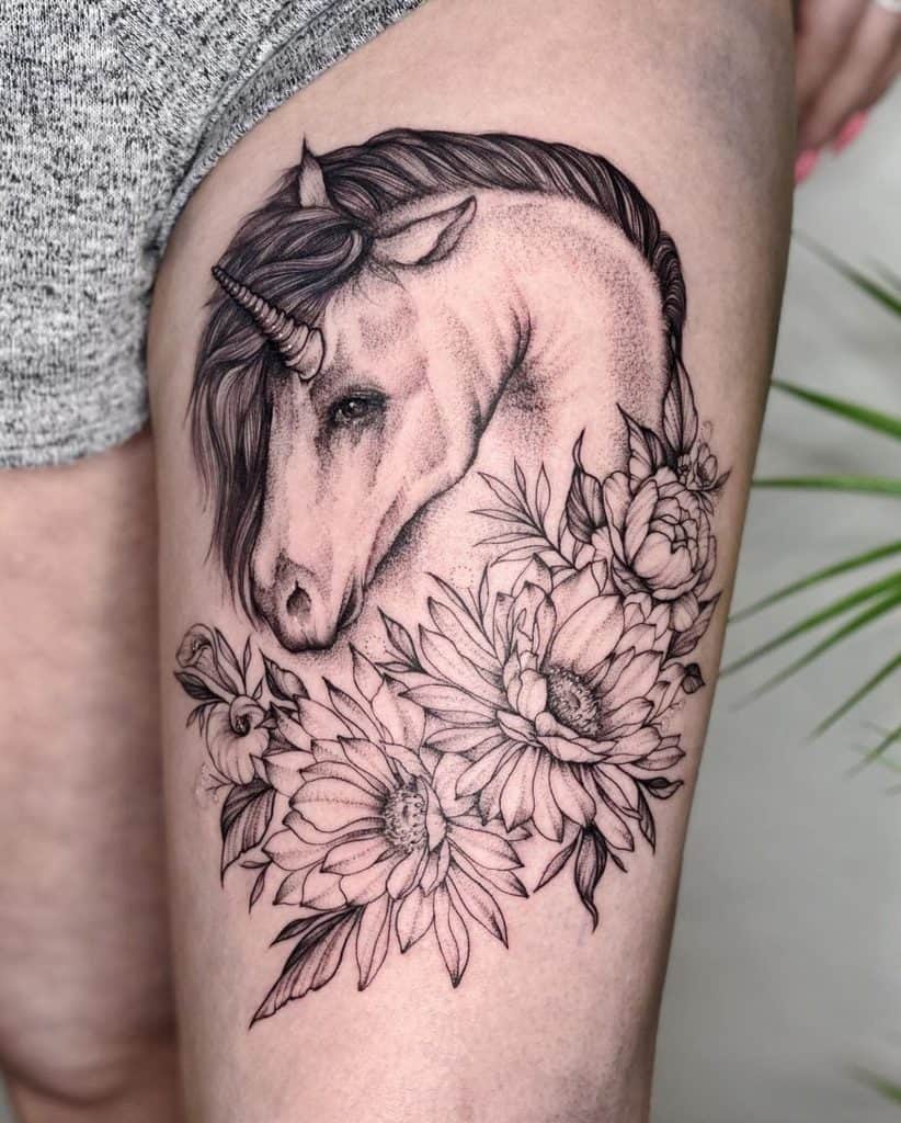 Elegant black and grey thigh tattoo of a unicorn with a braided mane, surrounded by detailed blooming flowers for a mystical, feminine look.