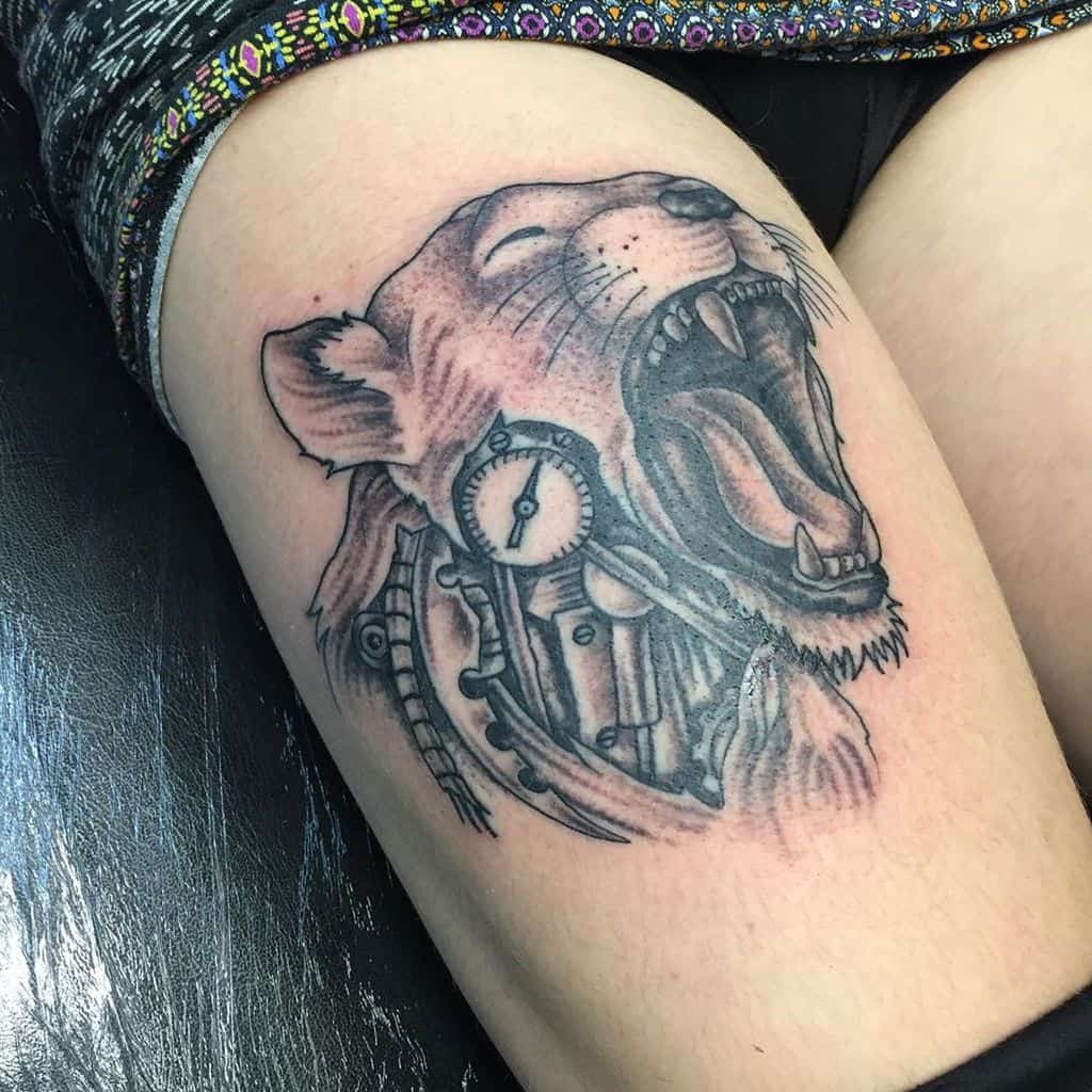 Steampunk-style roaring lioness tattoo on the thigh, featuring mechanical gears, pipes, and a clock integrated into the design.