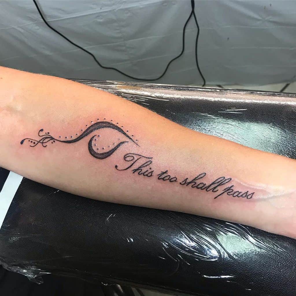 A tattoo on the forearm featuring the phrase "This too shall pass," with elegant curving lines and dot accents.