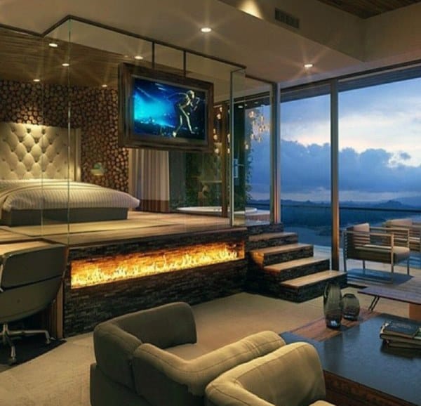 Luxurious bedroom with glass fireplace, elevated bed, and stunning sunset views through large windows.