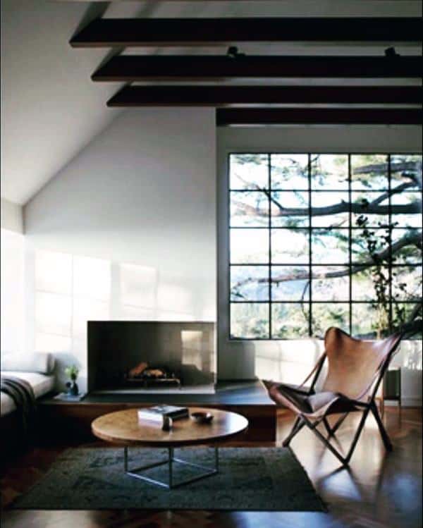 Modern living room with corner fireplace, large window, leather chair, and dark ceiling beams.