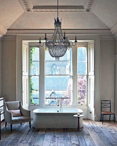 Rustic bathroom with a chandelier, bathtub, and large window overlooking trees and buildings
