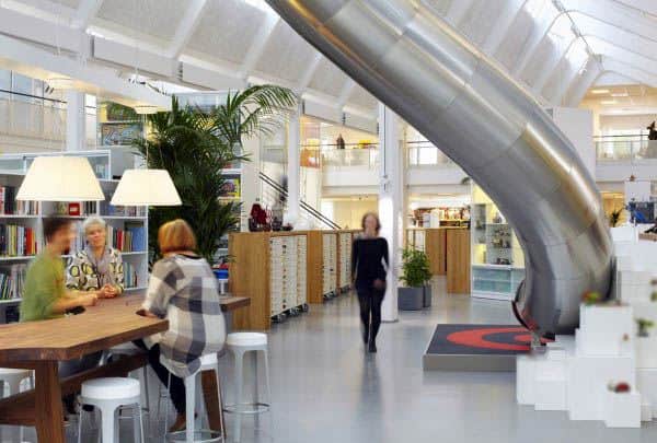Modern office with a slide, where people sit at a table and a woman walks by in the bright, open space