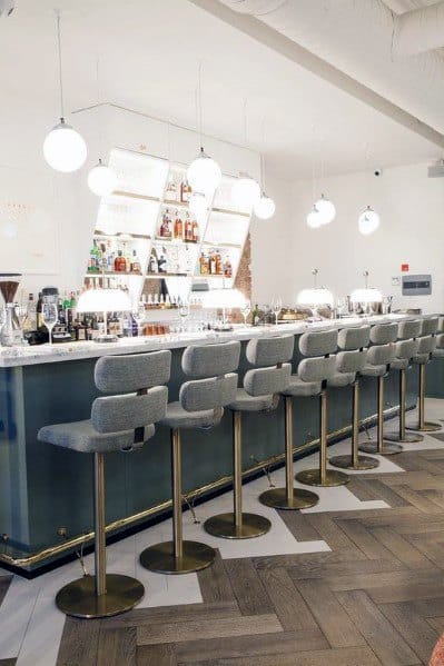 Modern bar with brass foot rail, upholstered grey barstools, and geometric shelving on a herringbone floor.