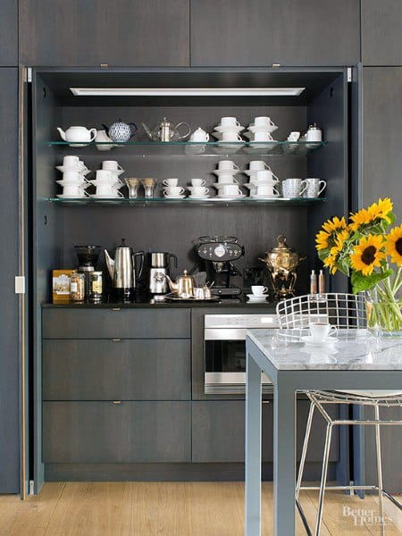 Dark built-in coffee bar with glass shelves, espresso machine, and a collection of teacups.