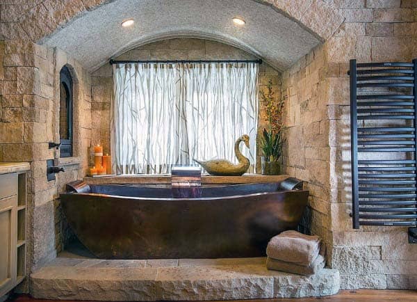 Rustic stone bathroom with a bronze tub, swan decor, and a towel on the floor