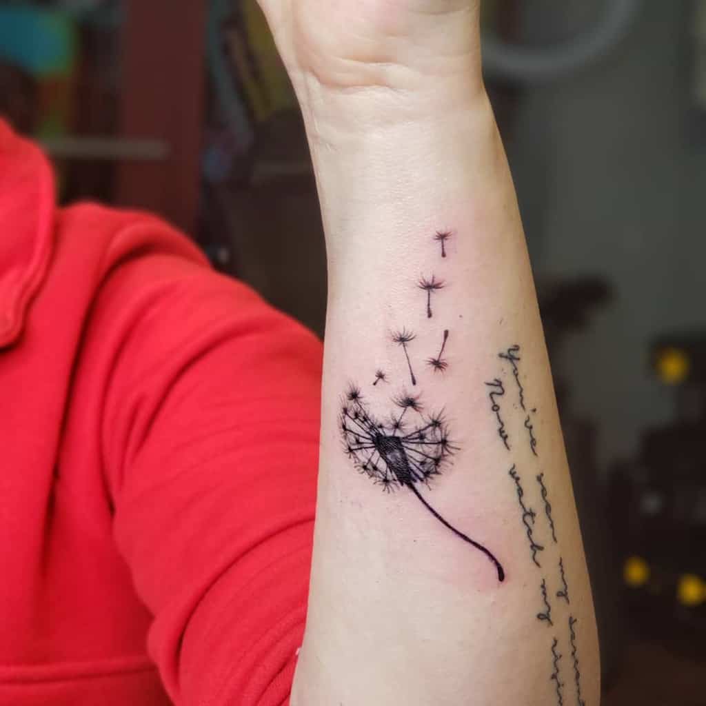 A dandelion tattoo on the forearm with seeds blowing off and the text "You're seeds of new reality" in delicate script beneath.