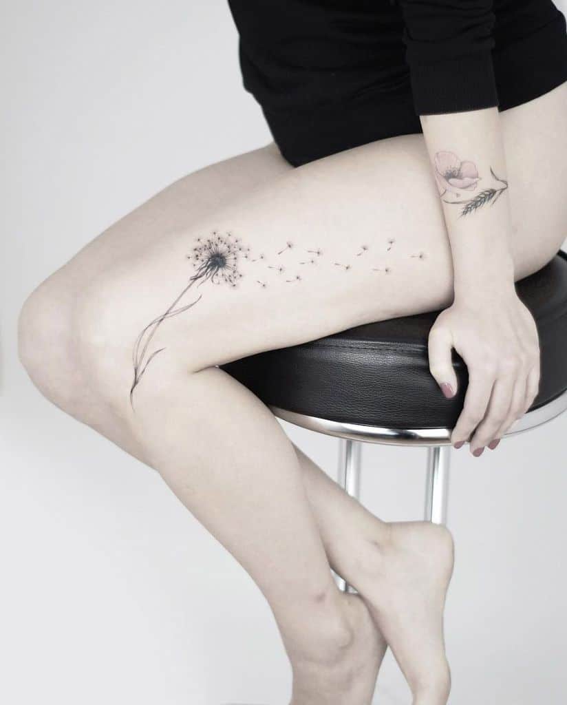 A delicate black and grey dandelion tattoo on the thigh with seeds floating away, complemented by a subtle floral tattoo on the arm.