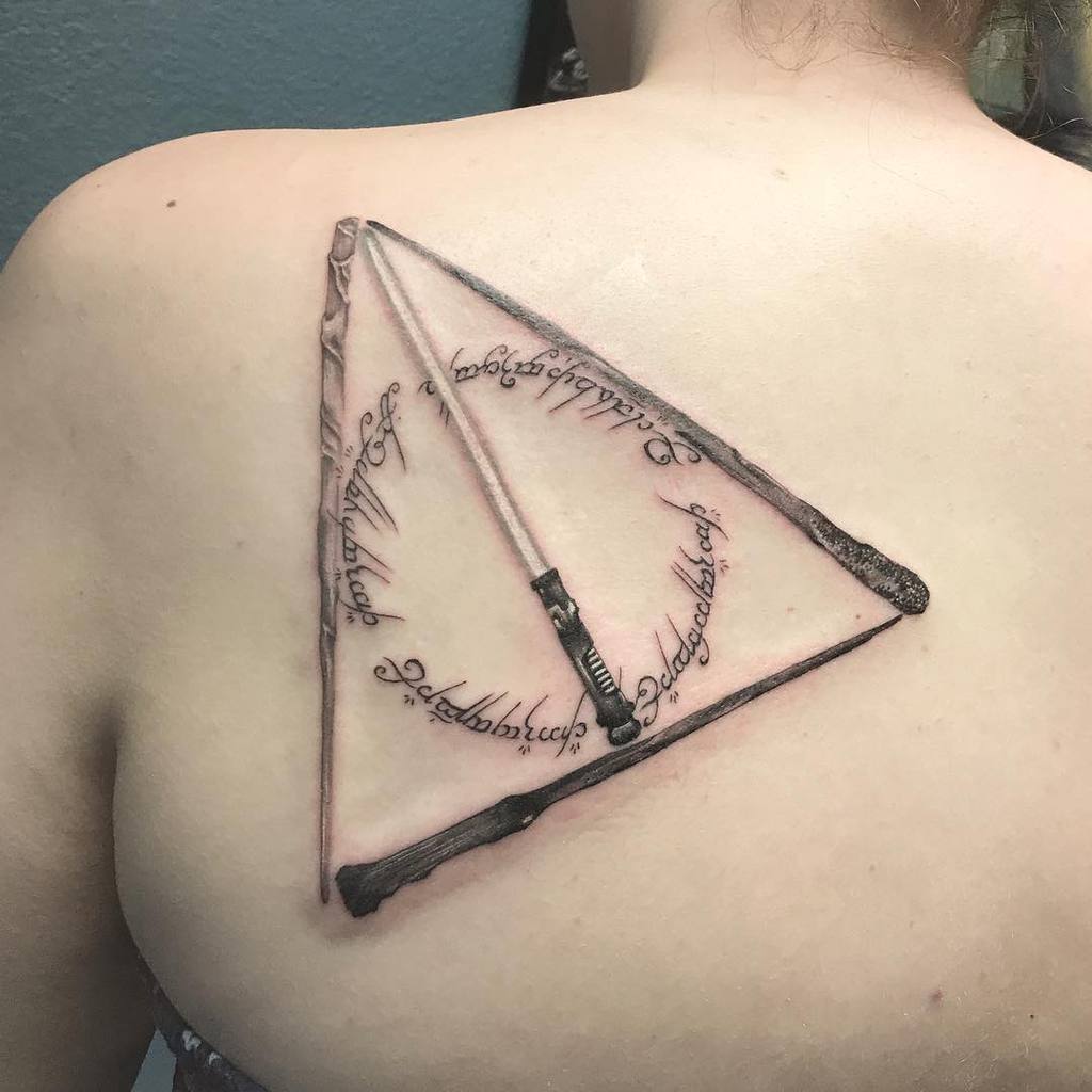 Realistic Deathly Hallows tattoo on the upper back, featuring wands forming the triangle with intricate script and a 3D skin-tearing effect.