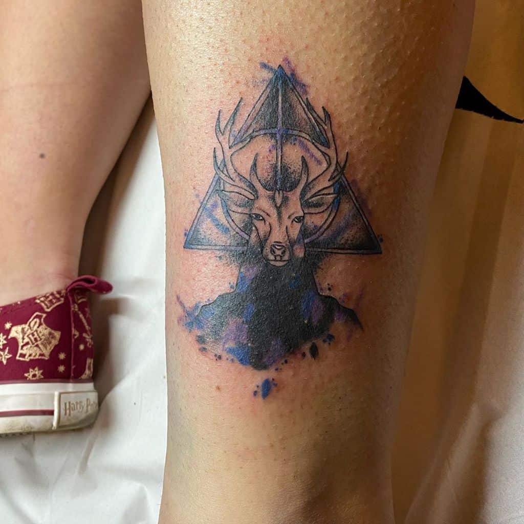 Deathly Hallows tattoo featuring a stag Patronus with antlers blending into the symbol, accented with watercolor-style blue and purple ink.