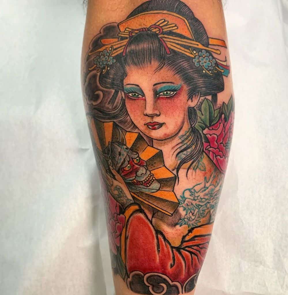 Colorful tattoo of a Geisha with bold makeup, intricate hair ornaments, holding a golden fan with a demon mask, surrounded by flowers.