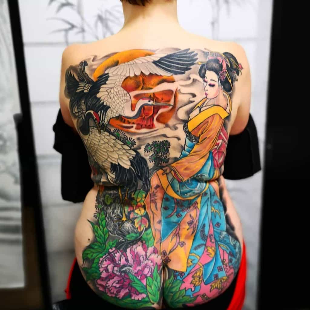 Vibrant full-back tattoo of a Geisha in a colorful kimono, with a crane, blooming flowers, and a sunset-lit pagoda in a traditional Japanese style.