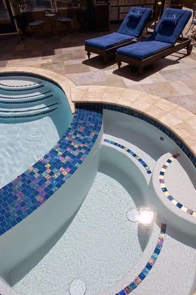 Curved pool and spa with blue iridescent mosaic tiles, surrounded by a stone patio and loungers.
