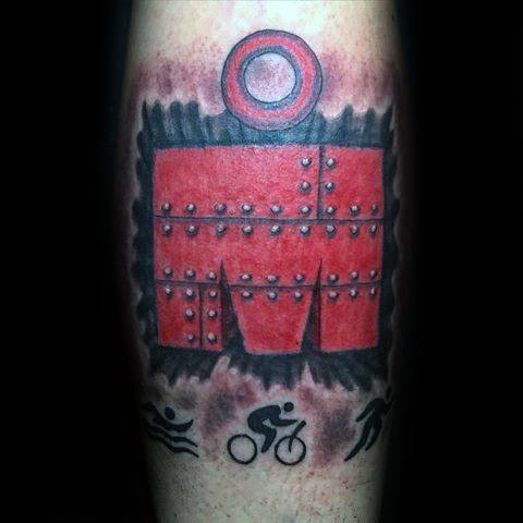 Ironman logo tattoo styled like a red metal plate with bolts, framed by a gear-like border and icons of swimming, biking, and running below.