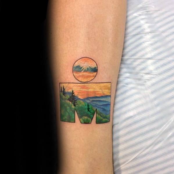 Ironman logo tattoo featuring a scenic mountain landscape with a colorful sunset sky, framed in a geometric design.