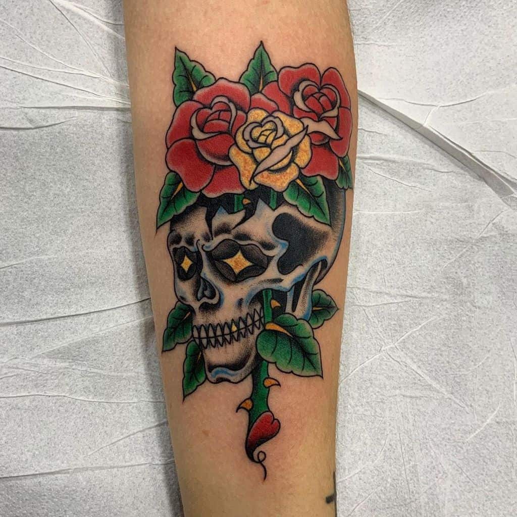 Neo-traditional tattoo of a skull with glowing eyes, surrounded by vibrant red and yellow roses and green vines, blending bold colors and shading.