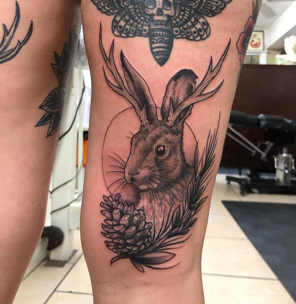 Intricate black and gray tattoo of a rabbit with antlers, framed by a pinecone and leaves, with circle line background