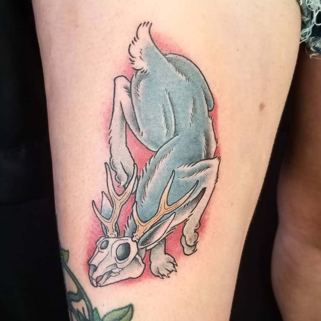 Stylized tattoo of a rabbit with antlers and a skull-like face, set against a soft red background, with detailed linework and shaded elements