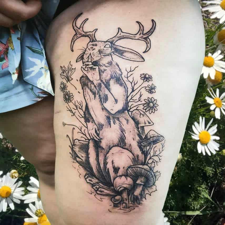 Intricate tattoo of a rabbit sitting in a wood with antlers, surrounded by wildflowers, mushrooms, and branches