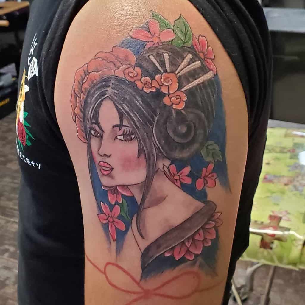Colorful upper arm tattoo of a Geisha with intricate floral hair ornaments, cherry blossoms, and a deep blue background for contrast.