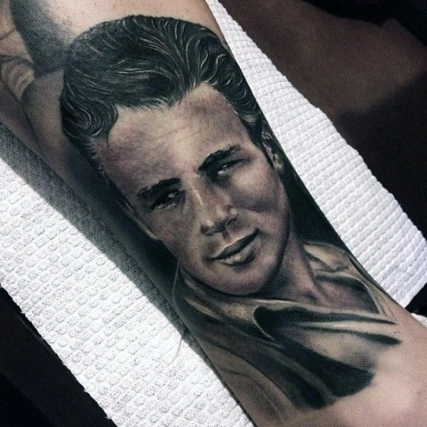 Portrait tattoo of James Dean with a confident smirk, detailed in black and grey with soft shading and classic attire.