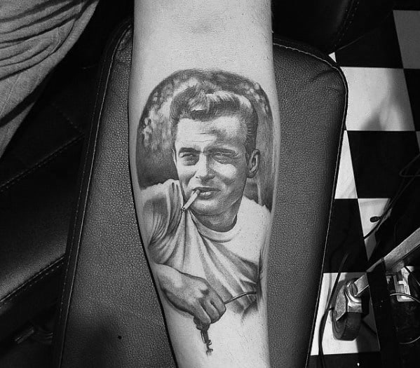 Black and grey calf tattoo of James Dean in a white tee, leaning casually with a cigarette in his mouth and a relaxed expression.