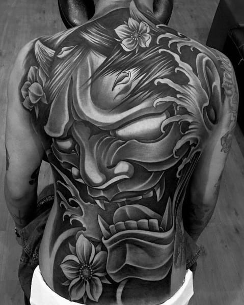 Large full-back tattoo in black and grey featuring a fierce Hannya mask with flowing waves and floral elements for a bold Japanese-inspired design.