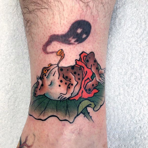 Japanese-style ankle tattoo featuring a relaxed frog lounging on a lotus leaf, smoking a pipe, with a playful ghostly spirit floating above.