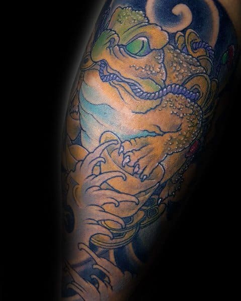 Japanese-style tattoo featuring a detailed golden toad gripping a scroll, surrounded by swirling waves and traditional elements, symbolizing wisdom and fortune.