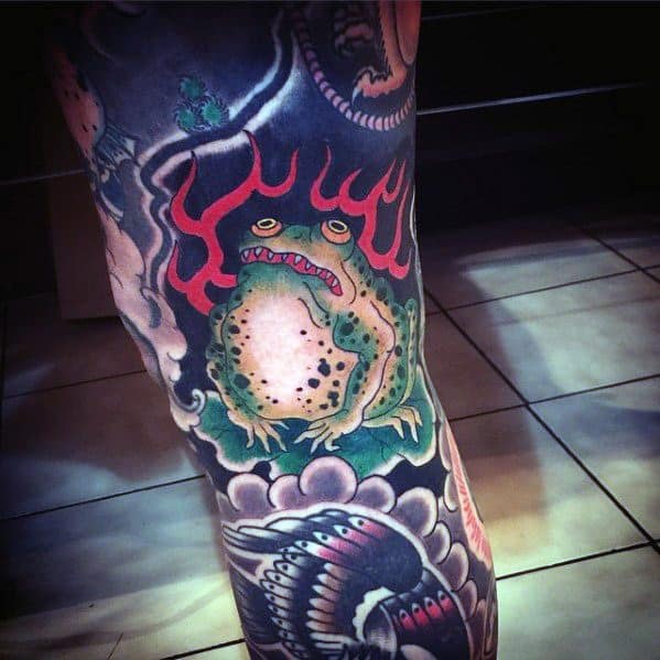 Japanese-style leg tattoo featuring a fierce green frog engulfed in red flames, surrounded by traditional swirling clouds and bold black ink patterns.
