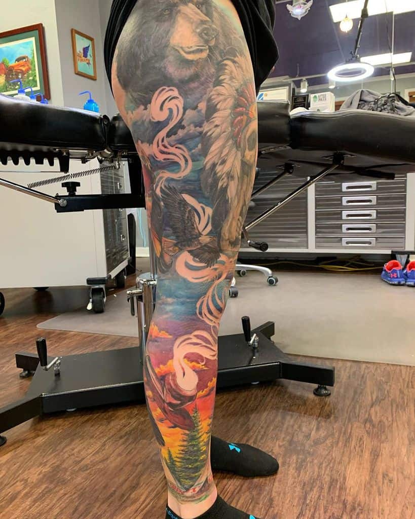 Leg sleeve tattoo featuring a bear, Native American headdress, eagle, smoke, colorful sky, clouds, cup on plate with smoke, pines and tents