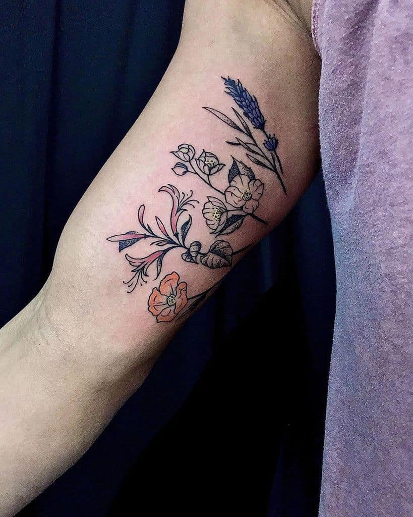 A tattoo of various flowers including lavender and other blossoms, with a mix of dark outlines and soft color accents, located on the upper arm.